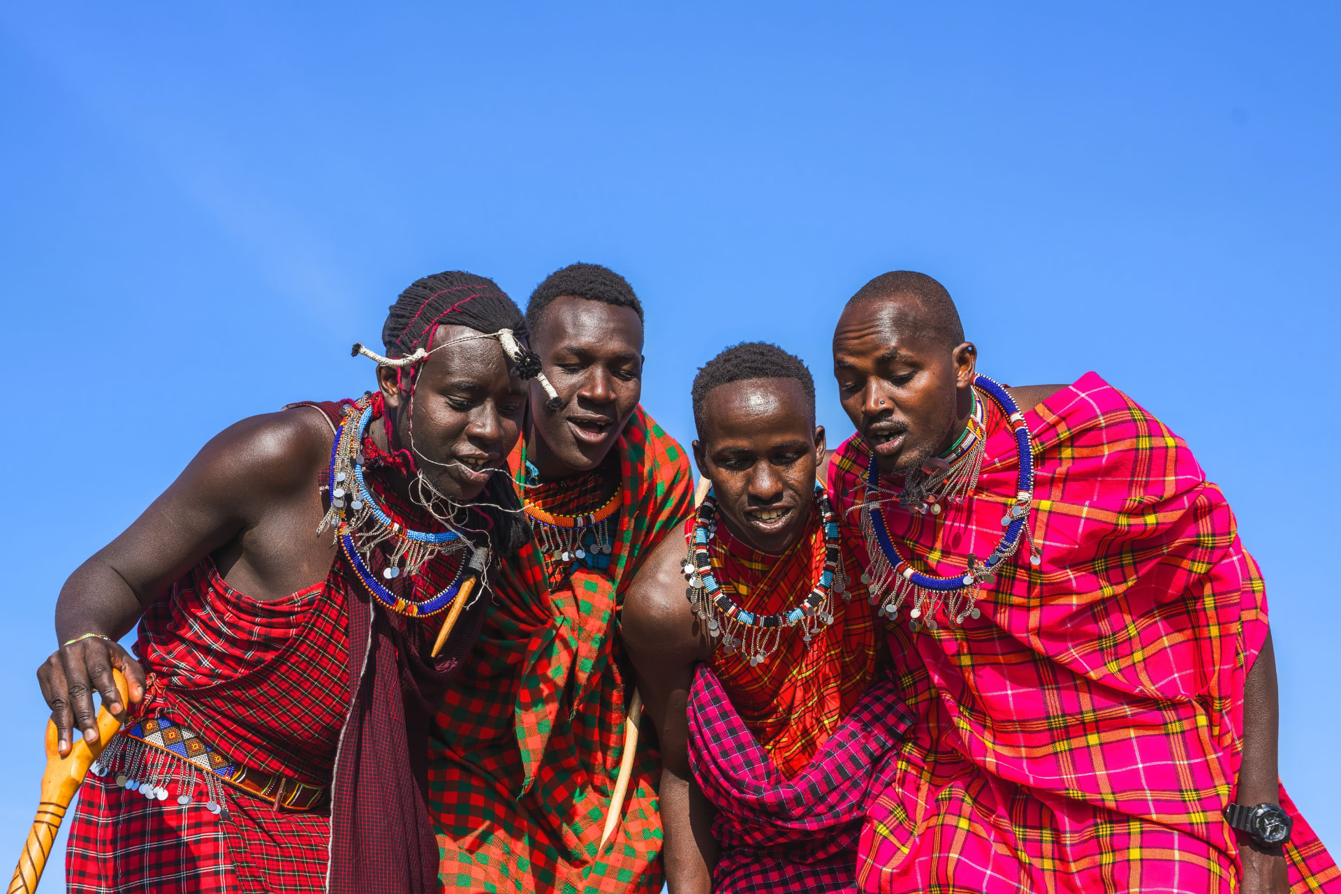 Maasai men in red and pink shukas singing together under a blue sky